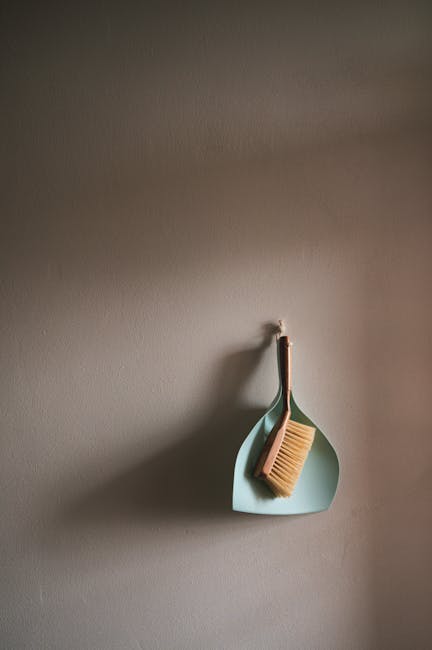 A light blue wall-mounted cleaning tool holder on a neutral wall, holding a wooden-handled scrub brush with beige bristles and a separate pale green dustpan. The brush is hanging from the holder, positioned slightly angled, with the dustpan nestled underneath. The lighting in the room is soft and even, highlighting the simple, clean surfaces and the minimalist arrangement, reflecting a focus on surface cleaning and maintenance as part of domestic cleaning services offered by Kingstonuponthamescarpetcleaners.com.
