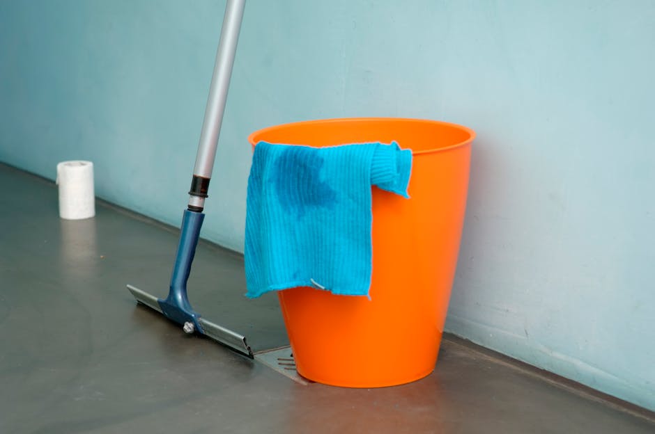 An indoor scene of a cleaning setup featuring a blue mop with a silver handle leaned against an orange plastic bucket positioned on a grey floor with a slight sheen. A blue cleaning cloth is draped over the edge of the bucket, appearing damp. In the background, a natural light illuminates a light-colored wall, while a roll of white paper towels sits on the floor nearby. The area suggests surface cleaning or deep cleaning activities in a domestic or commercial setting, with a focus on maintaining hygiene and sanitation, consistent with services offered by kingstonuponthamescarpetcleaners.com.