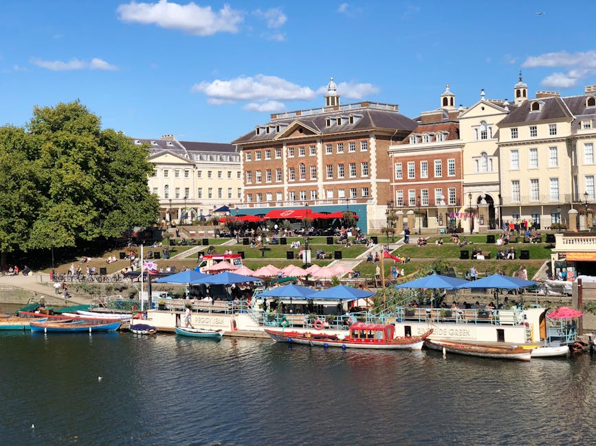 A riverside scene in Kingston upon Thames showing a row of moored boats with blue, pink, and red awnings along the water's edge. Behind the boats, there is a sloped grassy area with numerous people sitting on benches and on the grass, some under large umbrellas. The background features a historic multi-story building with a brick facade, white window frames, and decorative chimneys, all bathed in bright sunlight under a blue sky with scattered white clouds. The scene captures a lively, well-maintained outdoor setting emphasizing cleanliness and maintenance in a vibrant community area.