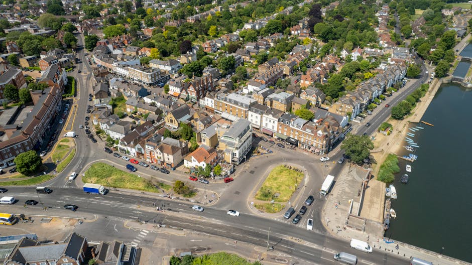 An aerial view of Kingston upon Thames showing a variety of buildings including commercial shops with glass windows and residential houses with tiled roofs. The streets are busy with cars, and there are small green spaces and trees lining the roads. To the right, a waterway with boats is visible, along with a promenade area. The image captures the mixture of urban infrastructure, greenery, and water, highlighting the well-maintained and clean environment typical of the area. Kingstonuponthamescarpetcleaners.com provides professional cleaning services that help maintain such pristine surfaces across homes and businesses.