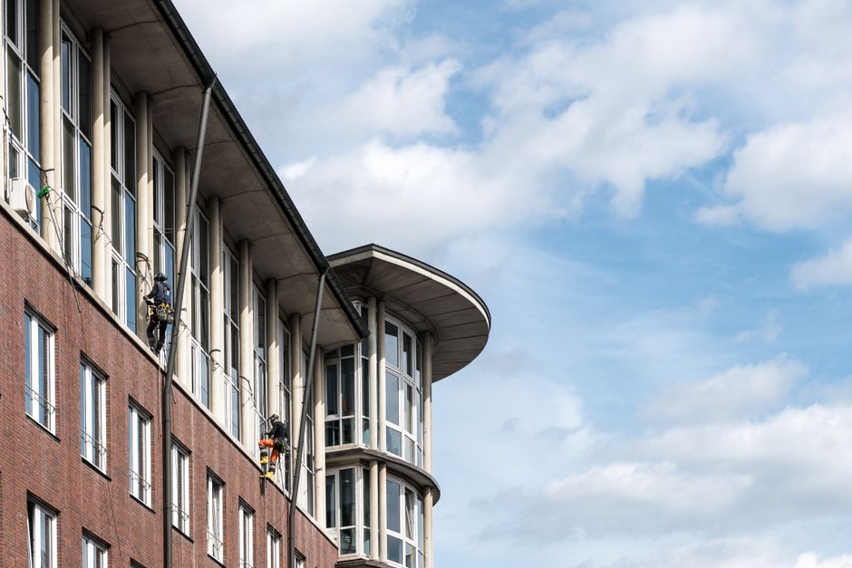 Exterior view of a modern multi-storey commercial office building with a brick facade and large glass windows, some of which are being cleaned by window washers suspended on ropes, using squeegees and cleaning tools. The building features a rounded corner with floor-to-ceiling windows, under a partly cloudy sky. The image highlights surface cleaning and maintenance in an urban environment, reflecting professional cleaning services offered by Kingstonuponthamescarpetcleaners.com for commercial properties like offices and business centers.