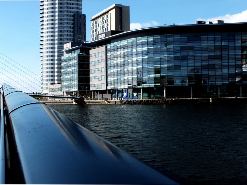 A modern commercial building with a predominantly glass facade reflecting the blue sky, located on Kingston Riverside Walk, with adjacent high-rise structures and a black river visible in the foreground. The building features large windows and a sleek design, illuminated by natural daylight, and the water below appears calm with no visible debris or dirt. The photograph captures the urban riverside scene, emphasizing cleanliness and architectural clarity typical of professional surface cleaning and maintenance standards, as promoted by Kingstonuponthamescarpetcleaners.com for business environments.