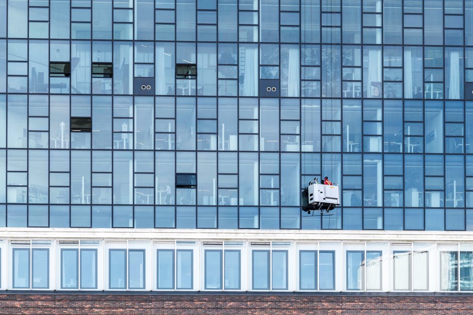 Exterior view of a modern multi-storey commercial office building with a brick facade and large glass windows, some of which are being cleaned by window washers suspended on ropes, using squeegees and cleaning tools. The building features a rounded corner with floor-to-ceiling windows, under a partly cloudy sky. The image highlights surface cleaning and maintenance in an urban environment, reflecting professional cleaning services offered by Kingstonuponthamescarpetcleaners.com for commercial properties like offices and business centers.