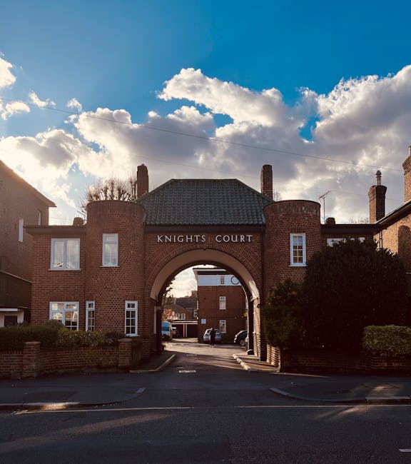 Photograph of the entrance archway to Knights Court on a bright day with partly cloudy sky. The brick arch is part of a residential building with brick walls, white window frames, and a tiled roof. The lane beneath the archway leads into a parking area with visible vehicles. Surrounding greenery includes bushes and trees on either side of the entrance. The scene is well-lit with natural sunlight, highlighting the clean and maintained exterior of the property, which may be associated with domestic or commercial cleaning services provided by KingstonUponThamesCarpetCleaners.com for end of tenancy cleaning near Kingston University.