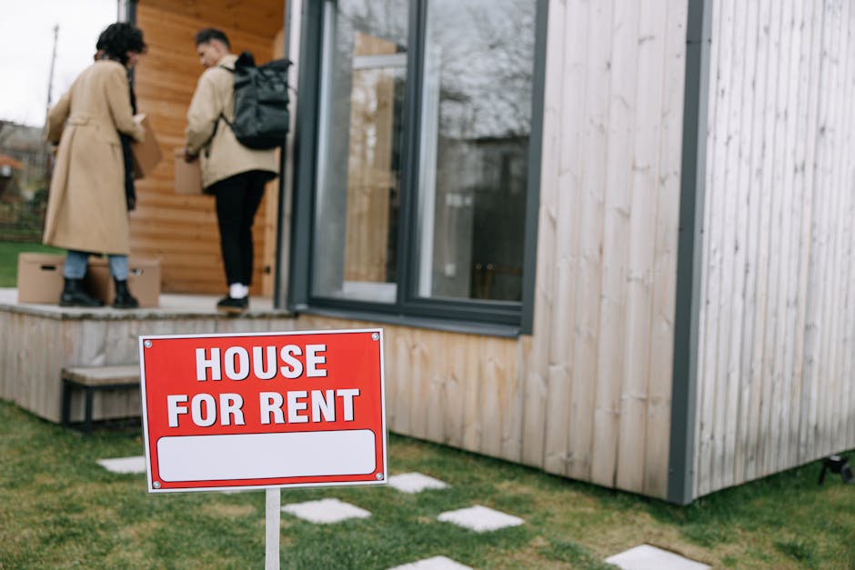 Exterior view of a wooden residential building with large glass sliding doors and a small wooden deck. Two people, dressed in beige and black clothing respectively, are standing on the deck engaged in conversation. A red and white 'House For Rent' sign is positioned in the foreground on a lawn with stepping stones. The building's surfaces include wooden siding and glass, with natural daylight illuminating the scene. The setting suggests a rental property prepared for new tenants, with a focus on maintaining clean, welcoming surfaces, consistent with end of tenancy cleaning services provided by Kingstonuponthamescarpetcleaners.com.