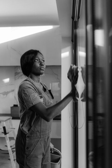 A professional cleaning expert from Kingstonuponthamescarpetcleaners.com is performing surface cleaning in a modern kitchen. The cleaner, an African woman wearing a grey uniform, is wiping a large glass door or window with a cloth, demonstrating detailed deep cleaning and sanitisation. The kitchen surface includes a stainless steel refrigerator, visible on the right, and a marble-effect backsplash on the wall behind her. The room is well-lit with natural light coming through the glass, highlighting the cleanliness and shine of the surfaces. The scene emphasizes precise cleaning processes, with no visible dust or streaks, showcasing the company's focus on thorough domestic cleaning and surface maintenance to ensure hygiene and a pristine environment.