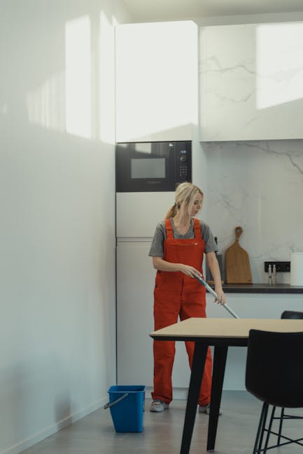 A professional cleaning expert from Kingstonuponthamescarpetcleaners.com is performing surface cleaning in a modern kitchen. The cleaner, an African woman wearing a grey uniform, is wiping a large glass door or window with a cloth, demonstrating detailed deep cleaning and sanitisation. The kitchen surface includes a stainless steel refrigerator, visible on the right, and a marble-effect backsplash on the wall behind her. The room is well-lit with natural light coming through the glass, highlighting the cleanliness and shine of the surfaces. The scene emphasizes precise cleaning processes, with no visible dust or streaks, showcasing the company's focus on thorough domestic cleaning and surface maintenance to ensure hygiene and a pristine environment.