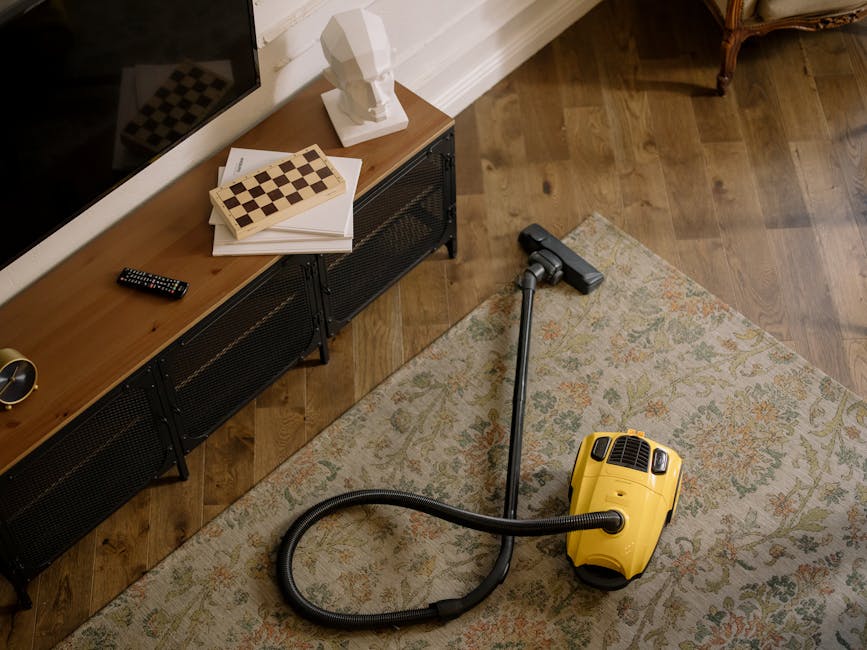 A yellow canister vacuum cleaner with a black hose and metal wand positioned on a traditional patterned area rug, in a living room with hardwood flooring, next to a wooden media unit with black metal legs. The media unit holds a remote control, a white bust sculpture, and a chessboard-shaped wooden box, all under warm ambient lighting. The room has white baseboards and crown molding, creating a clean and tidy appearance for surface cleaning and deep cleaning purposes, as promoted by Kingston upon Thames carpet cleaners.