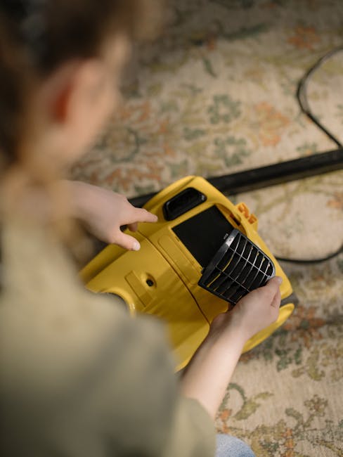 A person operating a yellow portable air mover or dehumidifier on a patterned carpet in a room with a textured, possibly brick or stone wall in the background. The individual is holding the device with both hands, focusing on its front grille. The carpet appears clean and dry, showcasing vibrant colors and a detailed floral or traditional design. Natural light illuminates the area, highlighting the cleanliness and maintenance of the surface. This scene is indicative of professional deep cleaning or surface drying processes by Kingstonuponthamescarpetcleaners.com, emphasizing hygiene and thoroughness in domestic cleaning services.