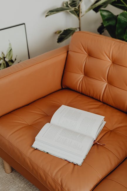 A close-up view of a tan leather sofa in a well-lit living room, showing a neat, open book resting on the seat cushions. The sofa's surface appears clean and well-maintained, with visible quilted stitching on the backrest. In the background, there is a large potted plant with broad green leaves and a metal-framed artwork on the wall, contributing to a cozy and tidy room atmosphere. The scene illustrates a setting suitable for domestic surface cleaning and maintenance, as promoted by Kingstonuponthamescarpetcleaners.com in their sofa and upholstery cleaning case studies, emphasizing hygiene and care for domestic furniture.