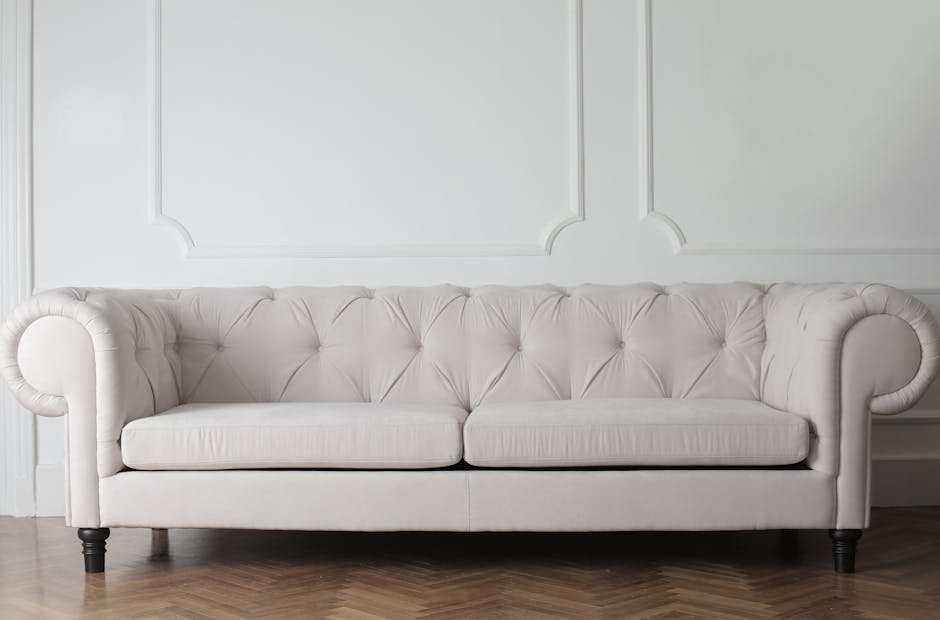 A clean, white, upholstered sofa with a tufted backrest and rolled armrests is positioned in front of a decorated white wall featuring molding details. The sofa's fabric appears freshly cleaned and well-maintained, with no visible stains or dirt. The dark wooden legs lift the sofa slightly above a light-colored wooden floor, which has a natural finish. The room is brightly lit, emphasizing the sofa's pristine condition and the neat appearance of the surrounding area, reflecting professional surface cleaning and deep cleaning processes in a domestic setting at Canbury, as part of a case study on sofa and upholstery cleaning by Kingston upon Thames Carpet Cleaners.