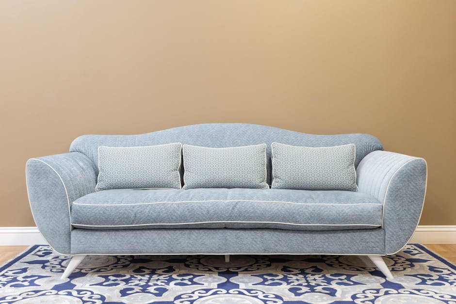 A close-up view of a tan leather sofa in a well-lit living room, showing a neat, open book resting on the seat cushions. The sofa's surface appears clean and well-maintained, with visible quilted stitching on the backrest. In the background, there is a large potted plant with broad green leaves and a metal-framed artwork on the wall, contributing to a cozy and tidy room atmosphere. The scene illustrates a setting suitable for domestic surface cleaning and maintenance, as promoted by Kingstonuponthamescarpetcleaners.com in their sofa and upholstery cleaning case studies, emphasizing hygiene and care for domestic furniture.