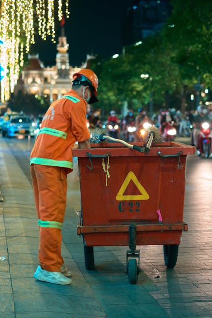 A sanitation worker dressed in an orange uniform, including a matching helmet and reflective safety strips, is seen collecting waste from a large red refuse bin on a city street during nighttime. The scene is illuminated by decorative string lights hanging from nearby trees, with historic buildings visible in the softly lit background. The worker is using a long-handled tool to lift trash from the bin, which appears to be placed on a paved sidewalk adjacent to a busy roadway filled with moving traffic. Nearby, green trees add a touch of nature to the urban environment. This scene emphasizes urban cleanliness efforts, with a focus on waste management and sanitation practices, consistent with information about disposal rules for cleaners in Kingston Council, as discussed on the page [PAGE_TITLE] on kingstonuponthamescarpetcleaners.com.