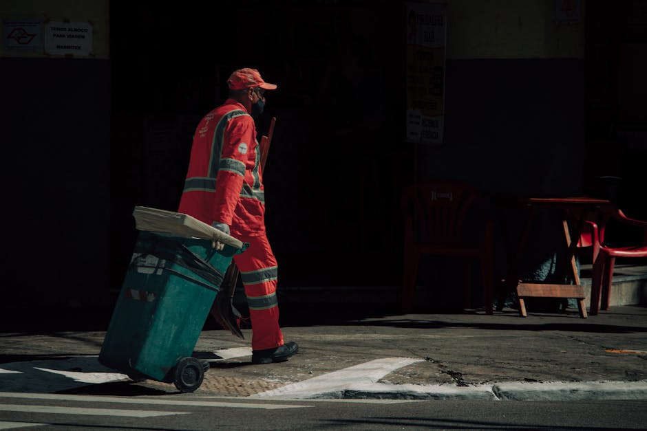 A sanitation worker dressed in a red uniform with reflective silver stripes is walking along a pavement, pushing a large green wheeled trash bin. The worker is wearing a cap and mask, and is illuminated by sunlight, creating a contrast with the darker background. The scene appears to be in front of a commercial or residential building, with red plastic chairs and a small table visible outside. The environment looks clean and well-maintained, emphasizing routine surface cleaning and waste disposal practices aligned with hygiene and sanitation standards, as noted on the page about disposal rules for cleaners Kingston Council at kingstonuponthamescarpetcleaners.com.