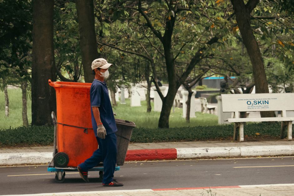A sanitation worker dressed in an orange uniform, including a matching helmet and reflective safety strips, is seen collecting waste from a large red refuse bin on a city street during nighttime. The scene is illuminated by decorative string lights hanging from nearby trees, with historic buildings visible in the softly lit background. The worker is using a long-handled tool to lift trash from the bin, which appears to be placed on a paved sidewalk adjacent to a busy roadway filled with moving traffic. Nearby, green trees add a touch of nature to the urban environment. This scene emphasizes urban cleanliness efforts, with a focus on waste management and sanitation practices, consistent with information about disposal rules for cleaners in Kingston Council, as discussed on the page [PAGE_TITLE] on kingstonuponthamescarpetcleaners.com.